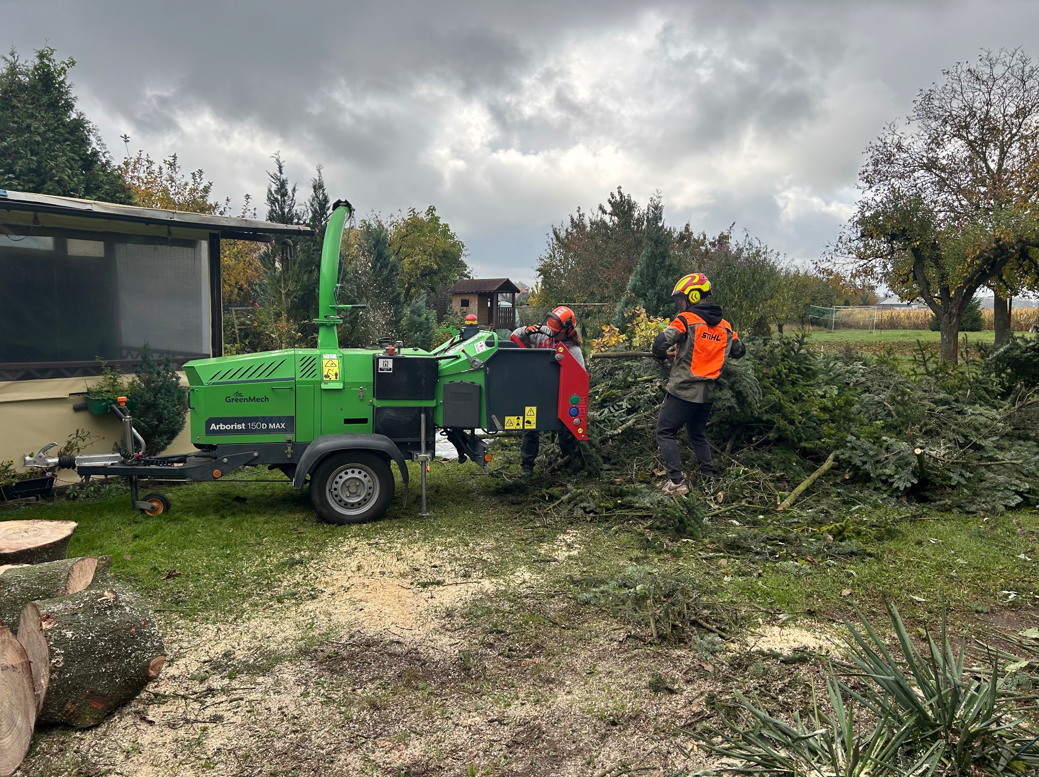 Pracownicy w kaskach wrzucają gałęzie do zielonego rębaka Arborist 150D MAX na tle ogrodu z domkiem. Wokół leżą pnie i wióry.