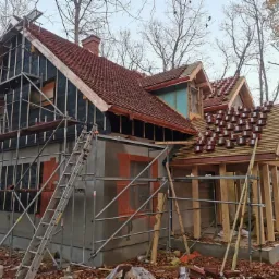 Dom w trakcie budowy, z widoczną więźbą dachową i częściowo ułożoną czerwoną dachówką, otoczony jesiennymi liśćmi i drzewami.