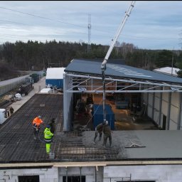 MaG Inwest - Wylewanie betonu na strop budynku w trakcie budowy, widoczna pompa do betonu, zbrojenie, pracownicy w odblaskowych ubraniach i częściowo zadaszona konstrukcja szkieletowa obok.