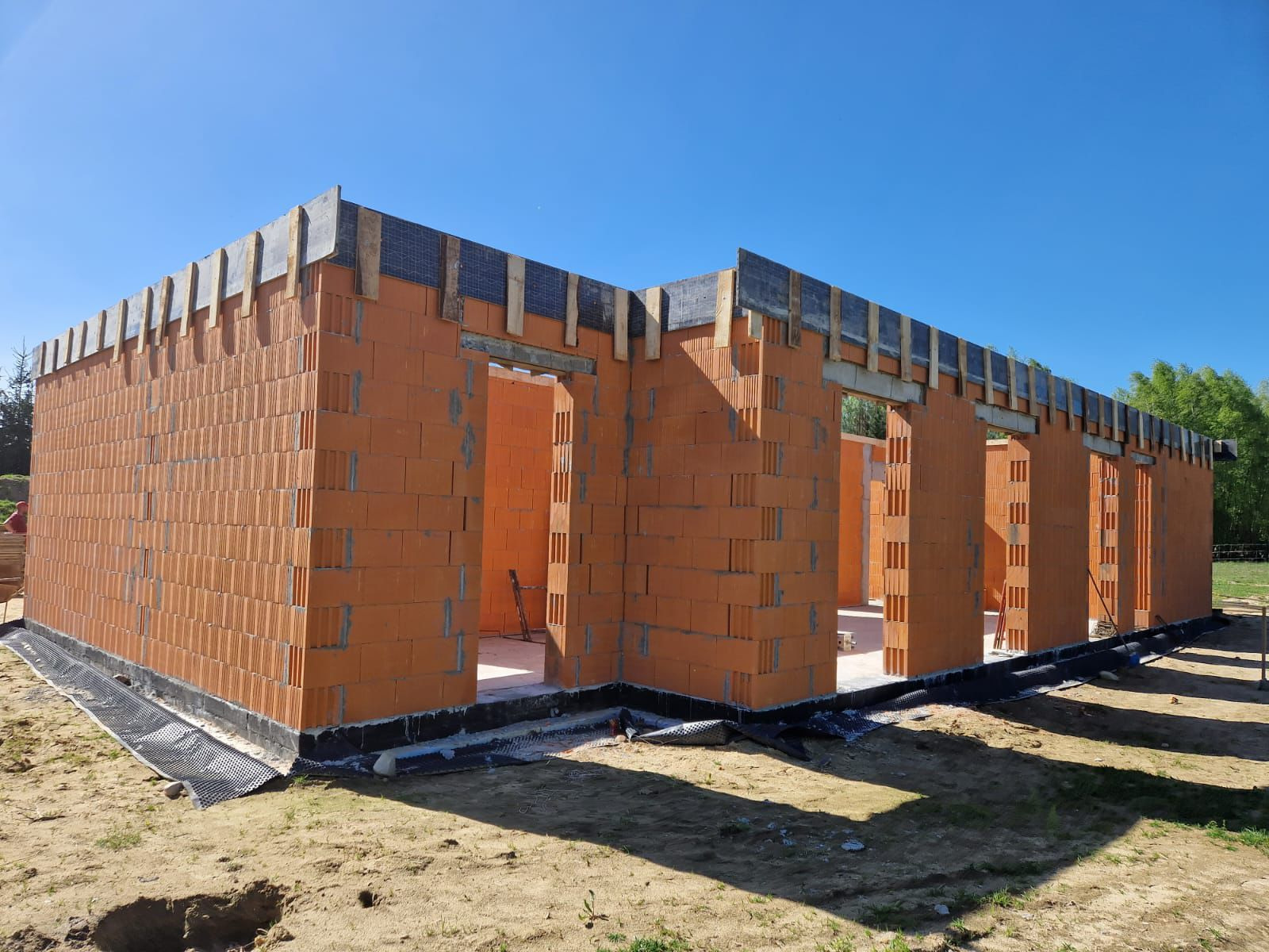 Budowa domu z czerwonej cegły ceramicznej Porotherm, widoczne ściany nośne, nadproża z drewna i folii, grunt wokół budynku.