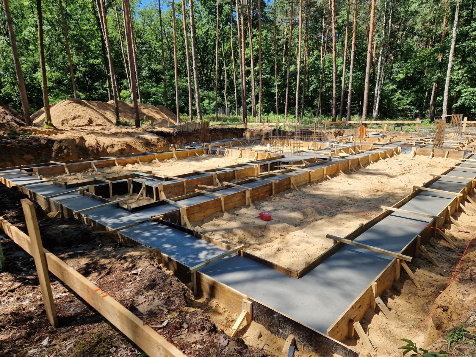 Fundamenty pod dom murowany w trakcie budowy w Warszawie, widok na betonową wylewkę w drewnianym szalunku i zbrojenie w tle, na piaszczystym terenie.