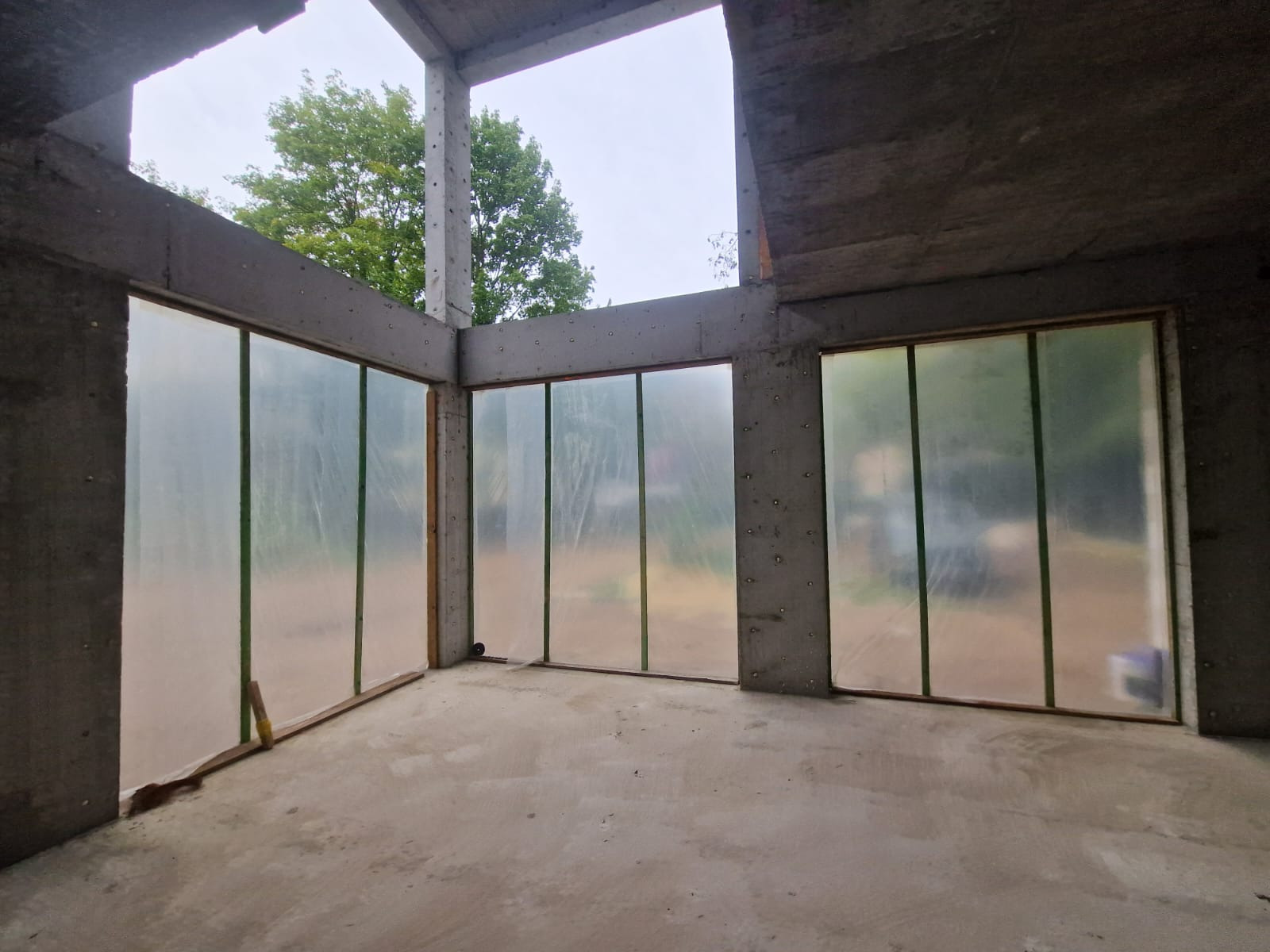 Surowy stan wnętrza domu w trakcie budowy z widocznymi betonowymi filarami i oknami zabezpieczonymi folią ochronną, widok na zieleń drzew przez otwory okienne.