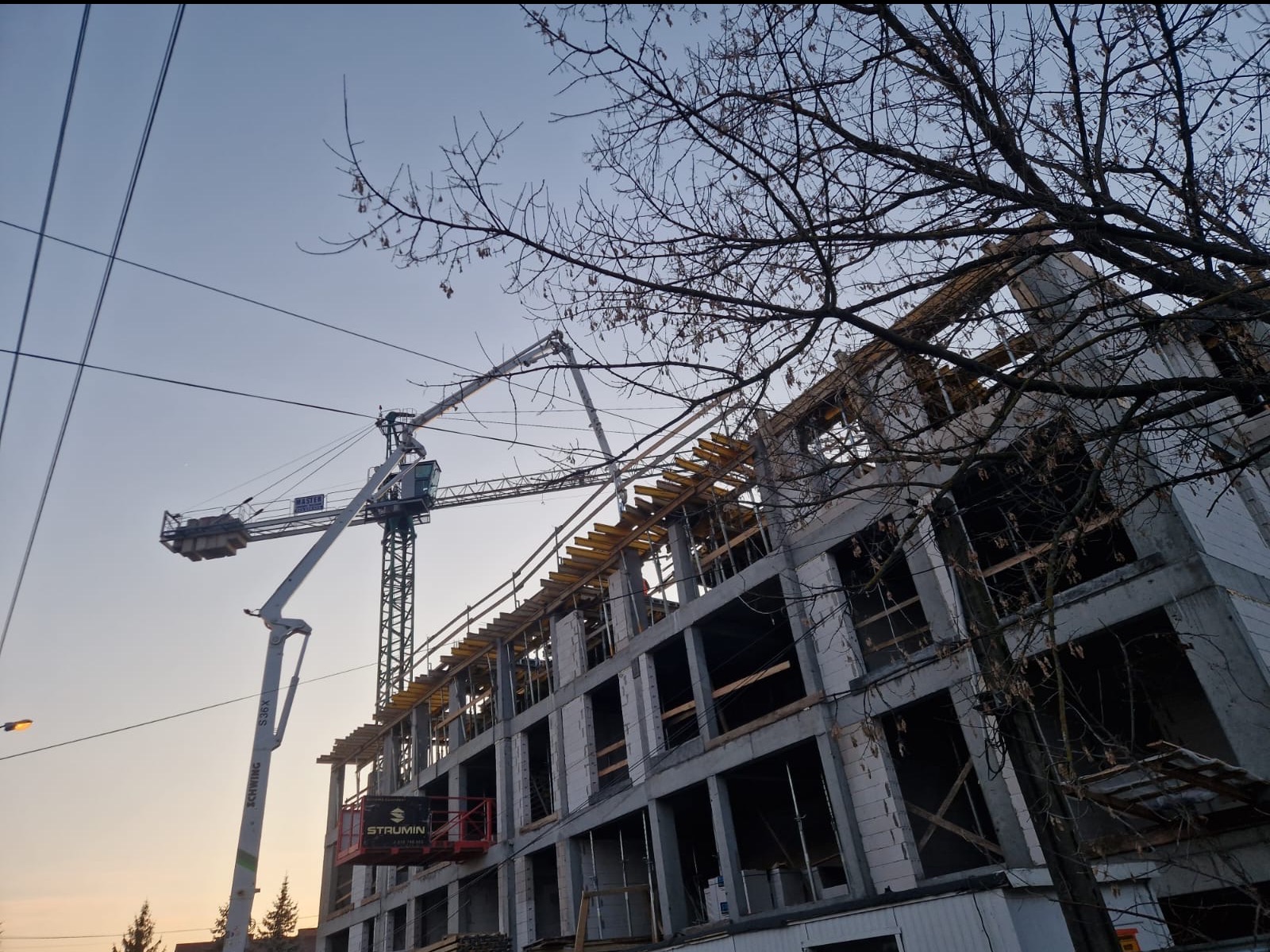 Wznoszenie wielopiętrowego budynku z betonu komórkowego w surowym stanie, widoczny żuraw budowlany z wysięgnikiem do podawania betonu i szalunki stropowe, na pierwszym planie gałęzie drzewa.