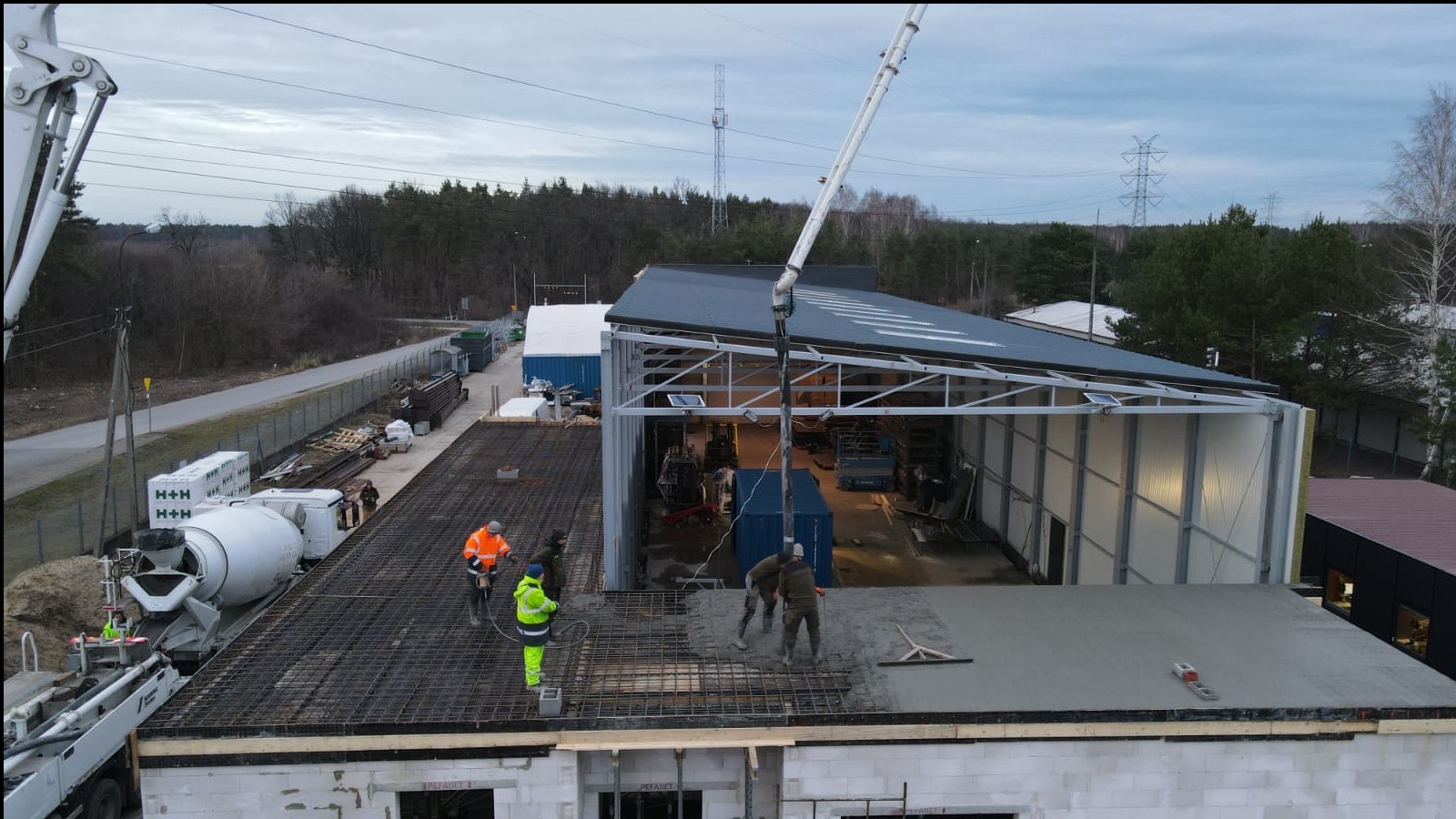 Wylewanie betonu na strop budynku w trakcie budowy, widoczna pompa do betonu, zbrojenie, pracownicy w odblaskowych ubraniach i częściowo zadaszona konstrukcja szkieletowa obok.