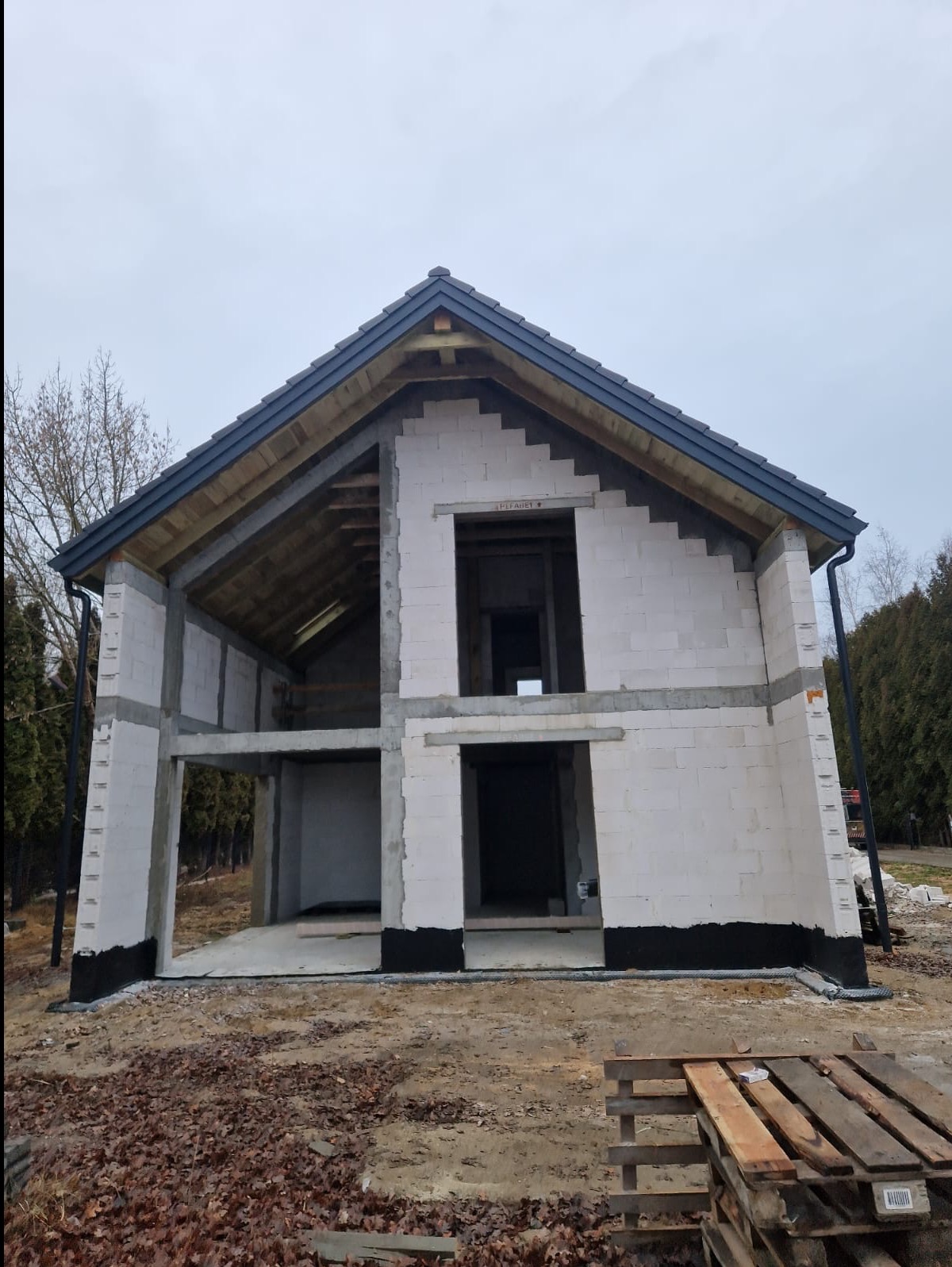 Surowy stan dwukondygnacyjnego domu z białego bloczka, widoczna więźba dachowa, czarne rynny, paleta na pierwszym planie, jesienne liście na ziemi.