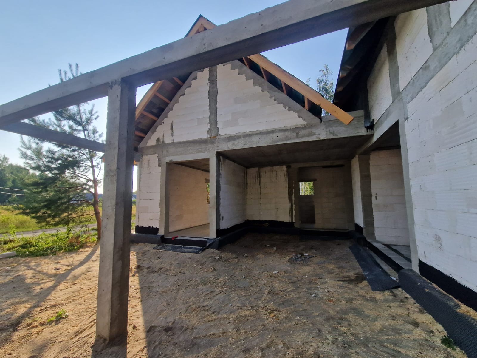 Surowy stan domu z betonu komórkowego w trakcie budowy, widoczne betonowe słupy i fragment więźby dachowej bez pokrycia, czarna izolacja przeciwwilgociowa przy fundamentach.