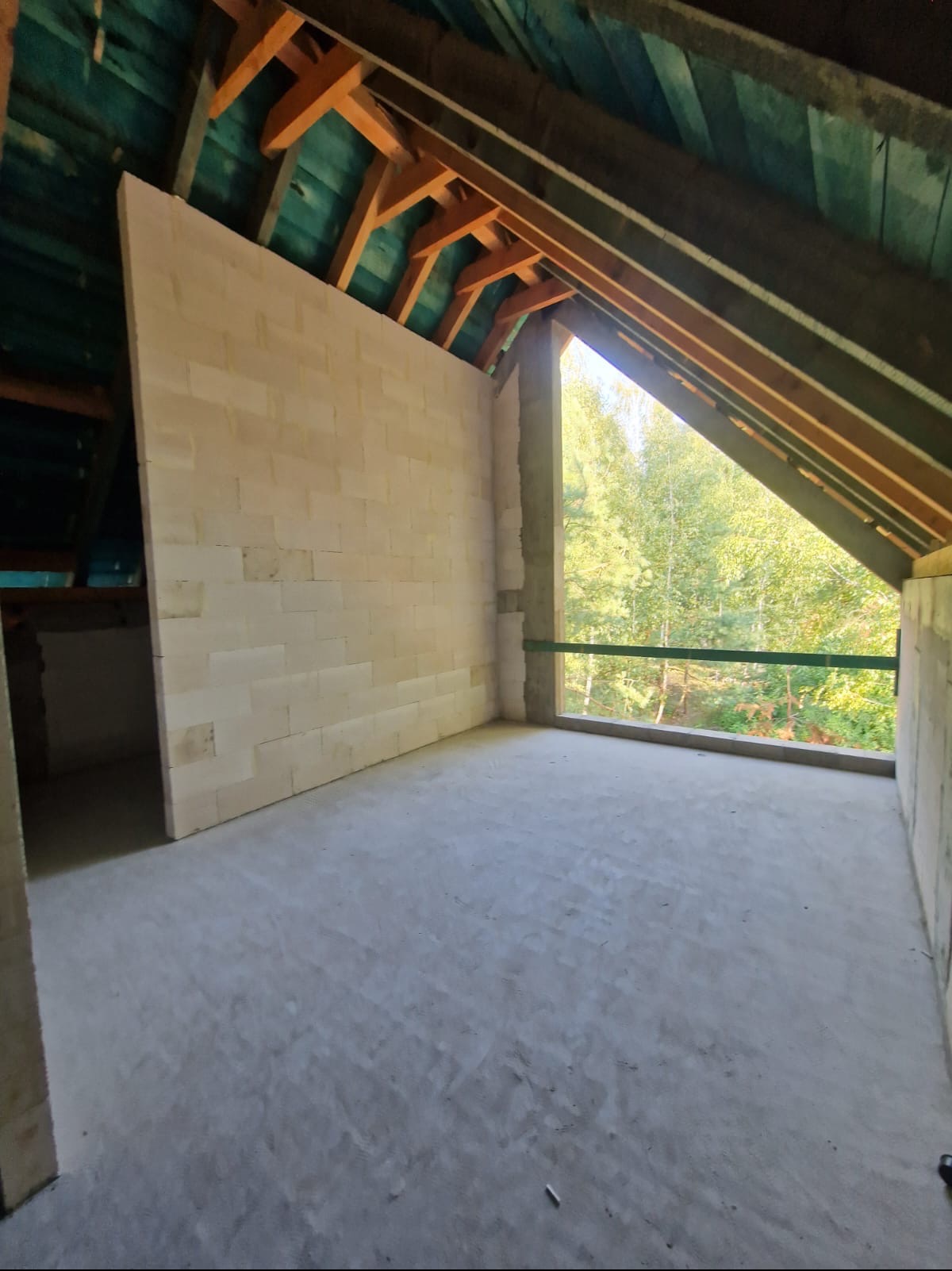 Wnętrze poddasza w trakcie budowy, ściany z białych bloczków, widoczna konstrukcja dachu z drewnianymi belkami, okno z widokiem na drzewa.