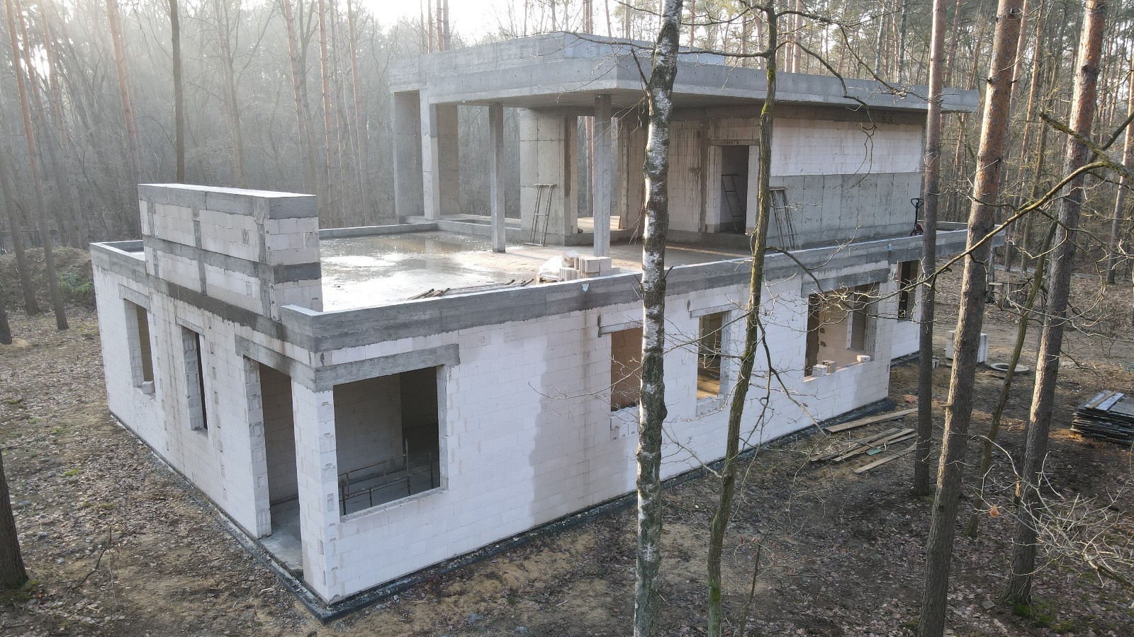 Niedokończony, dwukondygnacyjny budynek z białych bloczków, z widoczną konstrukcją dachu i otworami okiennymi, otoczony drzewami w lesie.