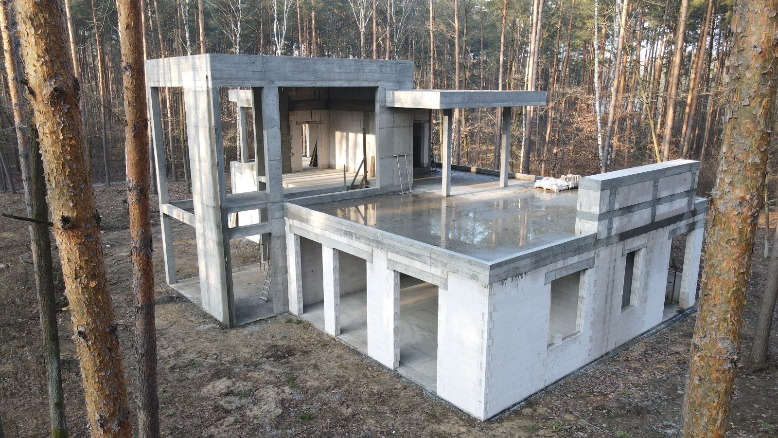 Surowy, dwukondygnacyjny dom z betonu komórkowego w trakcie budowy, z widocznymi elementami konstrukcyjnymi i mokrą powierzchnią dachu, otoczony lasem.
