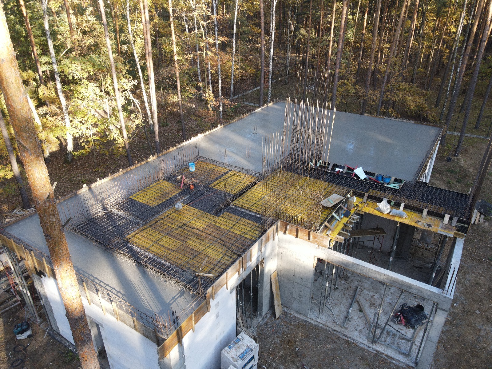 Wysoki kąt widzenia na fundamenty domu w trakcie budowy, widoczne zbrojenia, szalunki i częściowo wylany beton, otoczone lasem.