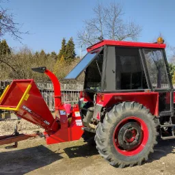 Czerwony traktor z zamontowanym rębakiem do gałęzi marki Remet CNC, stojący na żwirowym podłożu w słoneczny dzień.