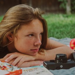 Adela Mróz - Młoda kobieta z piegami leży na kocu w plenerze, zamyślona, obok leżą jabłka, książka z motywem kwiatowym i stary aparat fotograficzny.