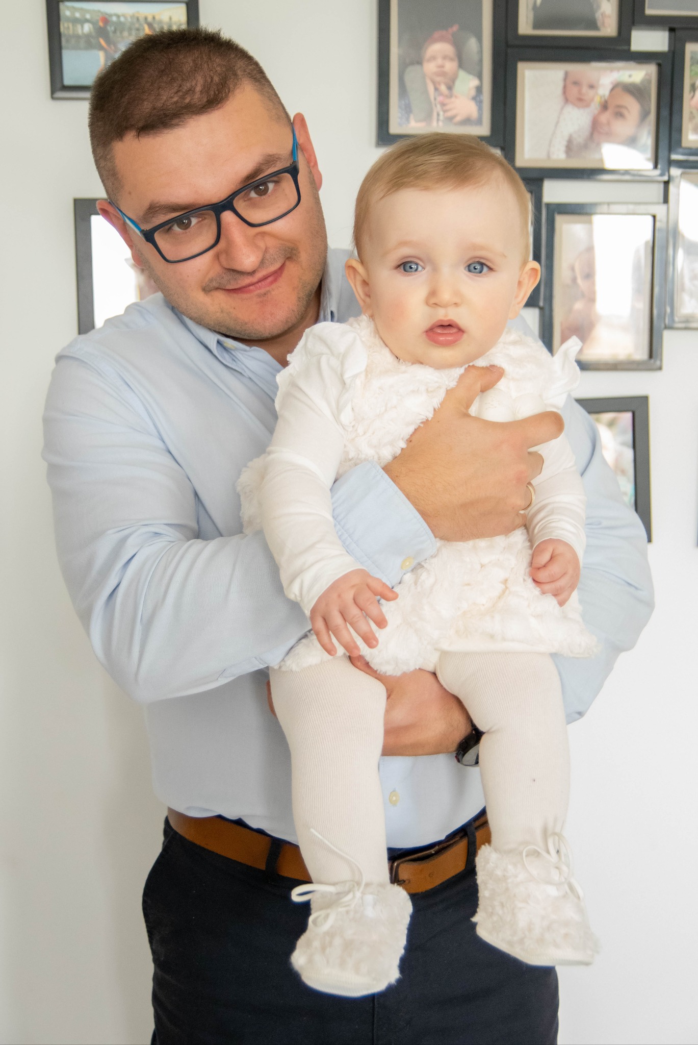 Ojciec w okularach trzyma na rękach niemowlę w białej sukience, w tle ściana z ramkami ze zdjęciami.