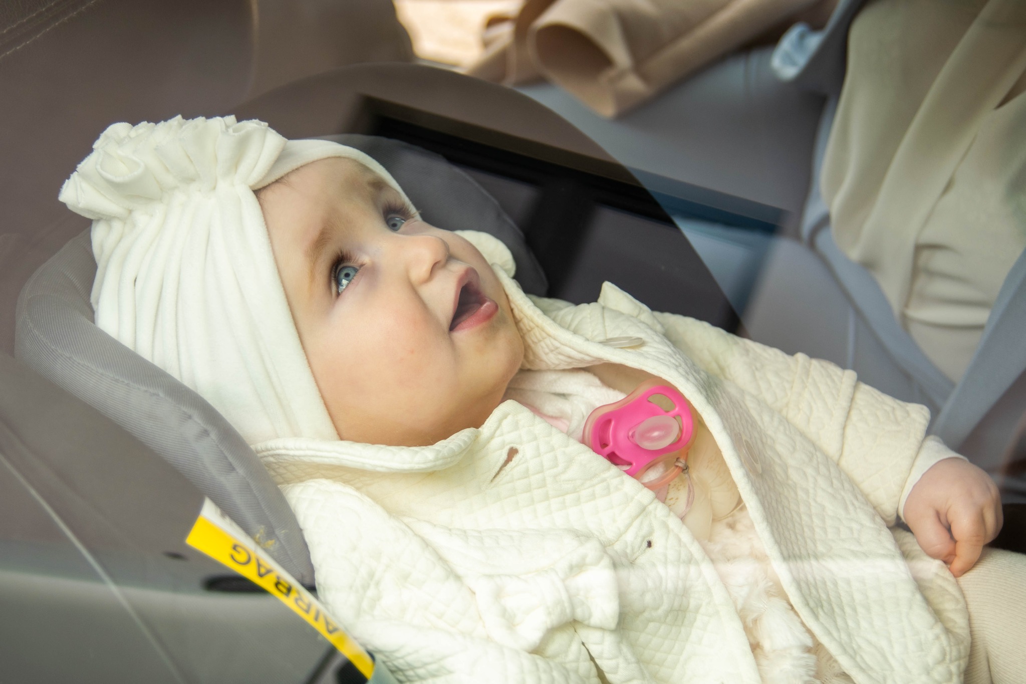 Niemowlę z niebieskimi oczami w białym ubranku i czapeczce, leżące w foteliku samochodowym z różowym smoczkiem w ustach, widziane przez szybę.