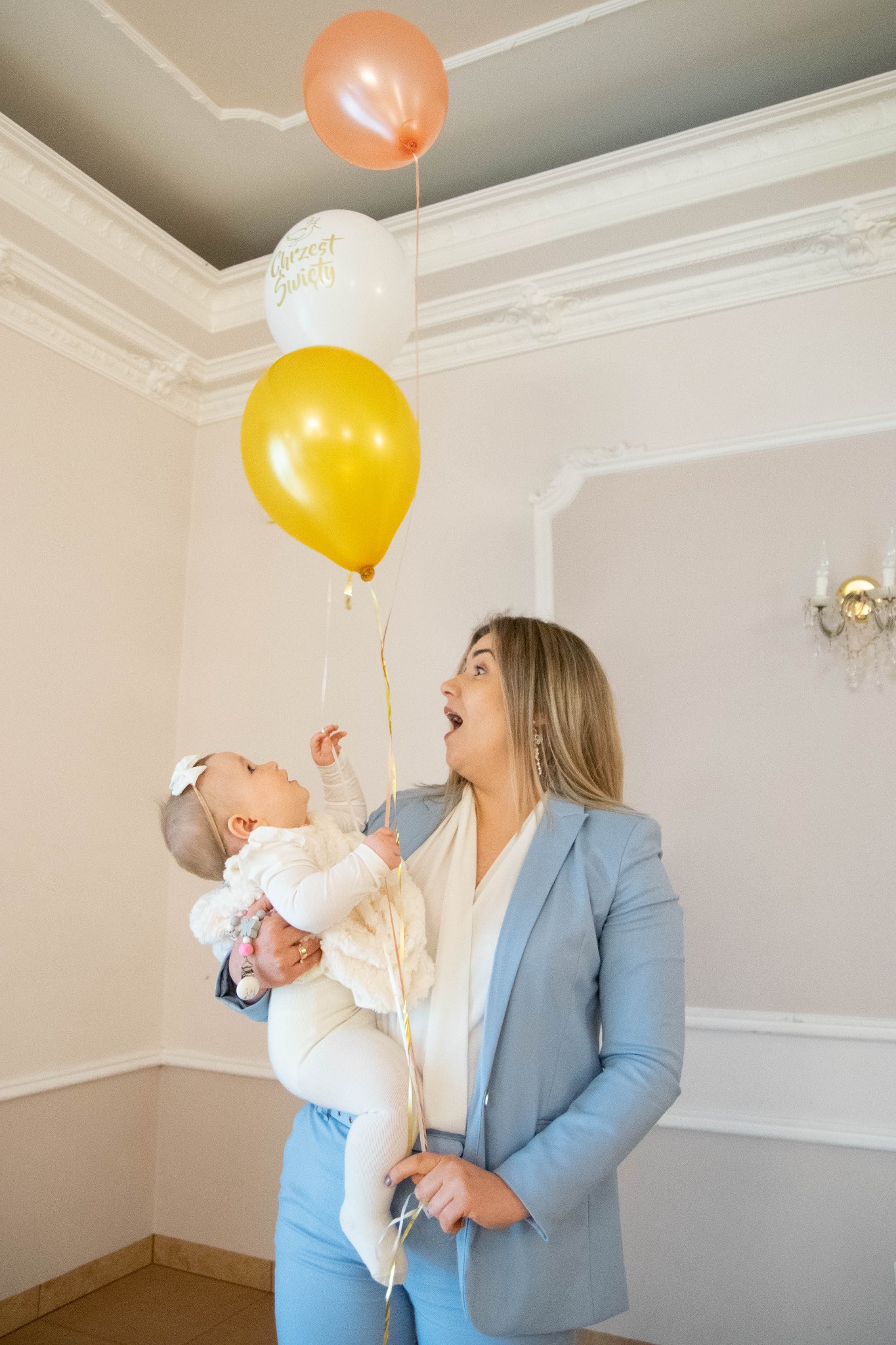 Kobieta w błękitnym garniturze trzyma na rękach niemowlę w białym ubranku, oboje spoglądają na balony w kolorach żółtym, białym z napisem 'Chrzest Święty' i brzoskwiniowym.
