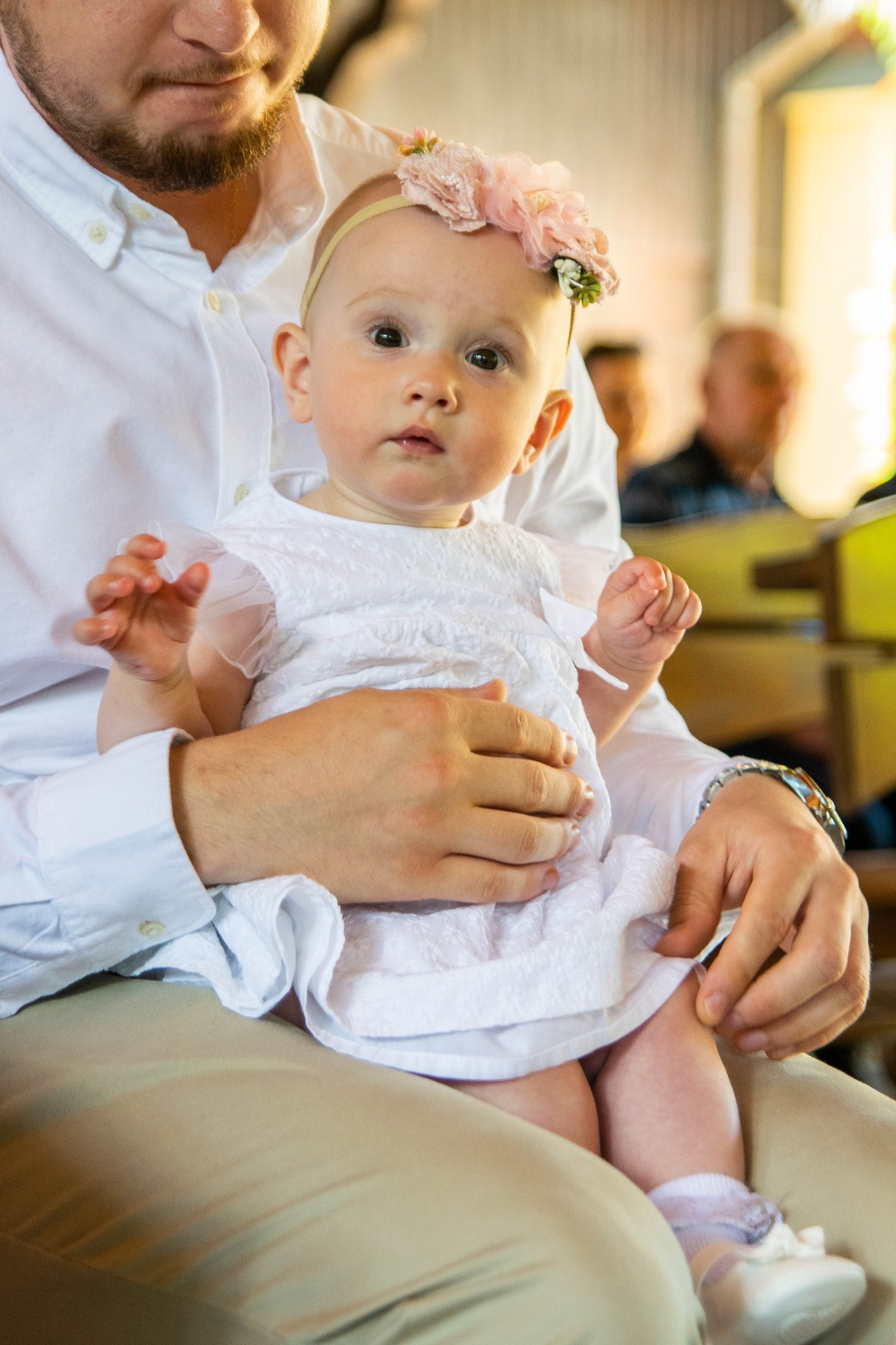 Urocza dziewczynka w białej sukience i kwiatowej opasce siedzi na kolanach u mężczyzny w jasnej koszuli, prawdopodobnie podczas uroczystości religijnej.