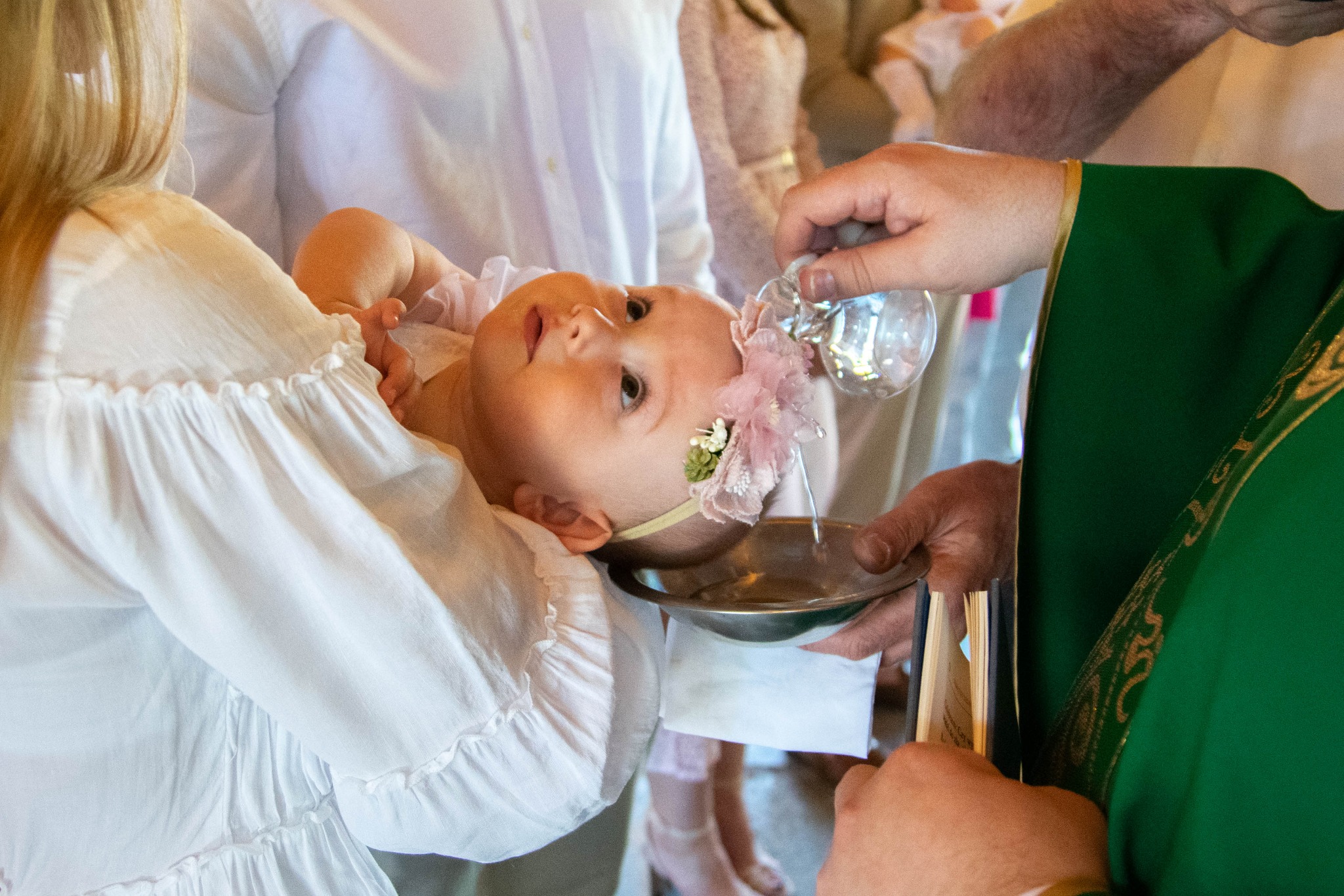 Ksiądz polewa wodą głowę niemowlęcia z kwiatową opaską podczas ceremonii.