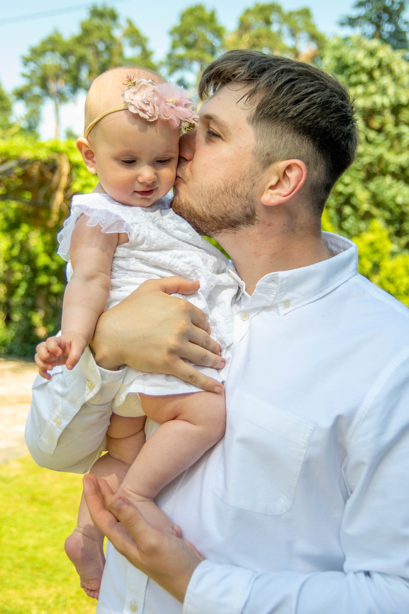 Tata w białej koszuli całuje w czoło małą dziewczynkę w białej sukience z kwiatową opaską na głowie, na tle zieleni.