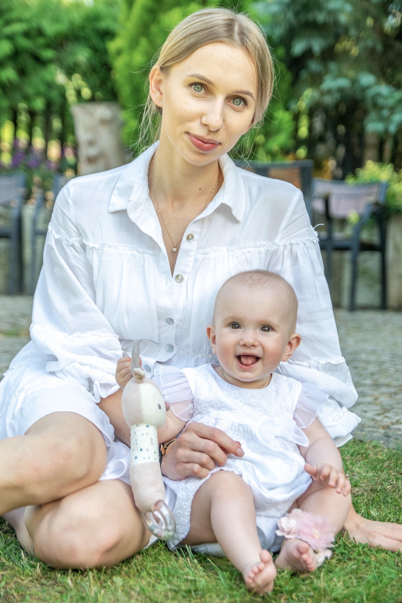 Portret matki z uśmiechniętą córeczką w białych sukienkach, siedzących na trawie w plenerze w Warszawie. Dziecko trzyma pluszową zabawkę.