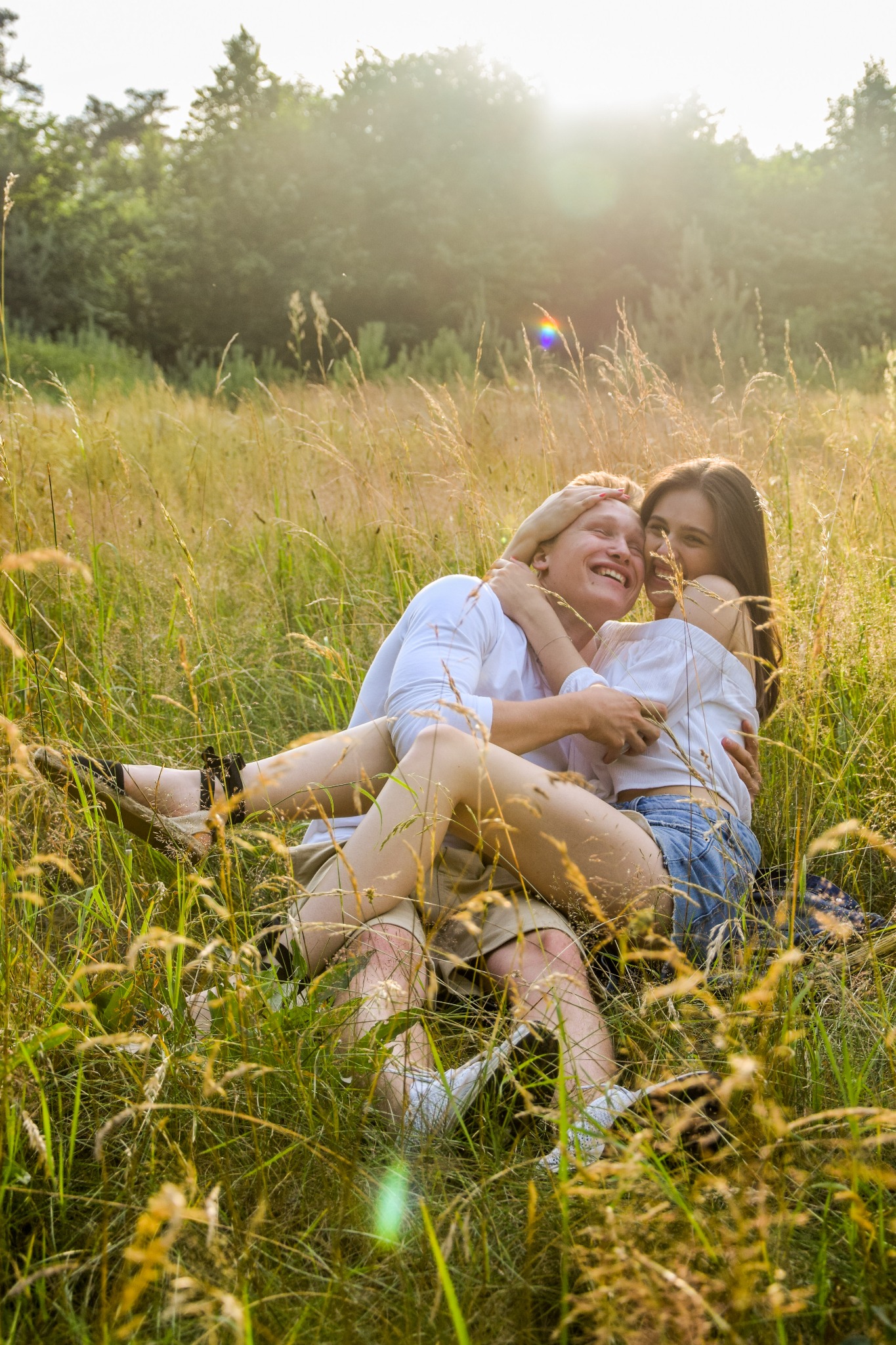Zakochana para w jasnych ubraniach przytulona w wysokiej trawie podczas sesji narzeczeńskiej w słoneczny dzień.
