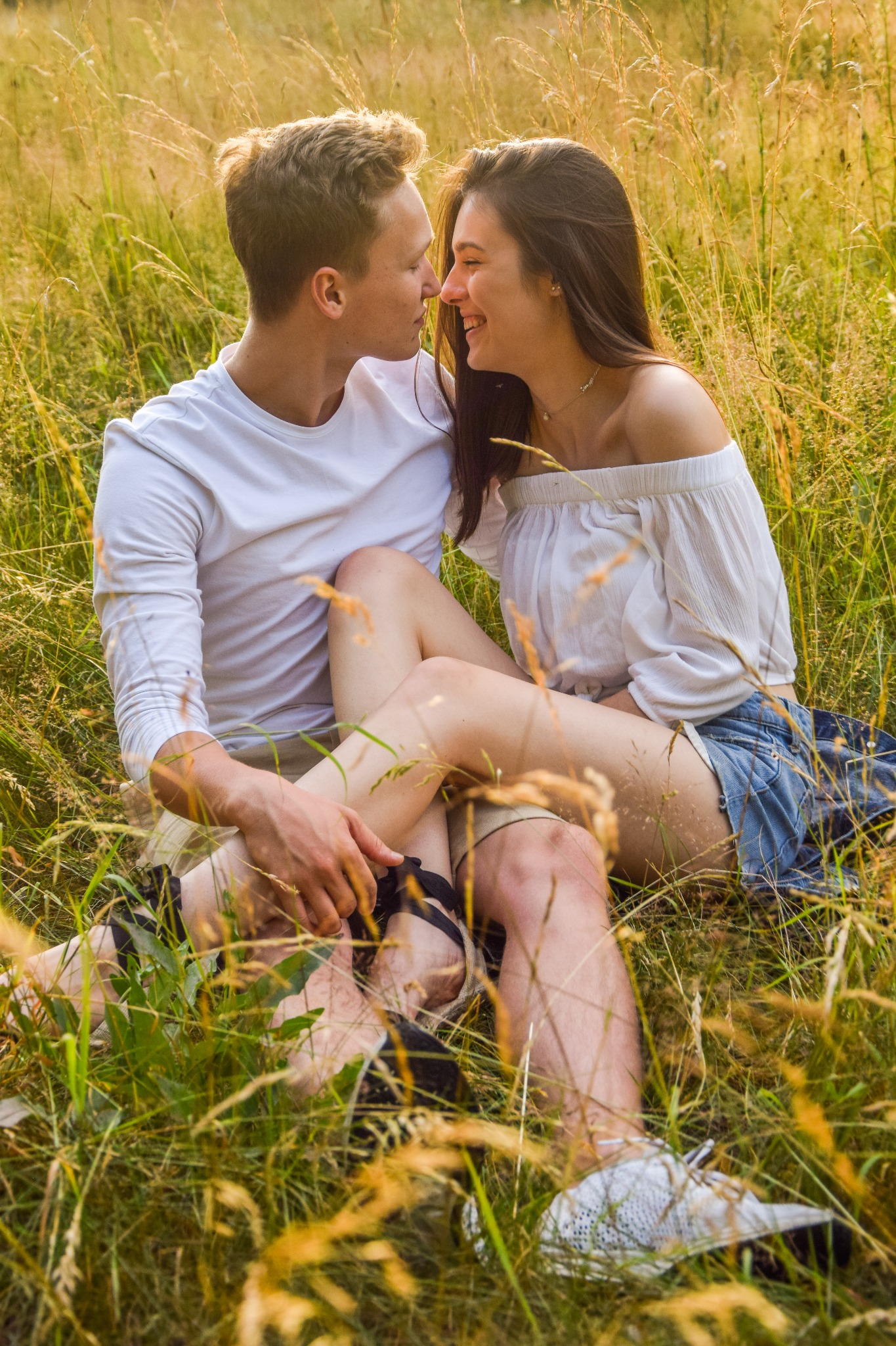 Uśmiechnięta para siedzi w wysokiej trawie podczas złotej godziny, bliskość podkreślona dotykiem nosów, ona w białej bluzce z odkrytymi ramionami i jeansowych szortach, on w białej koszulce...