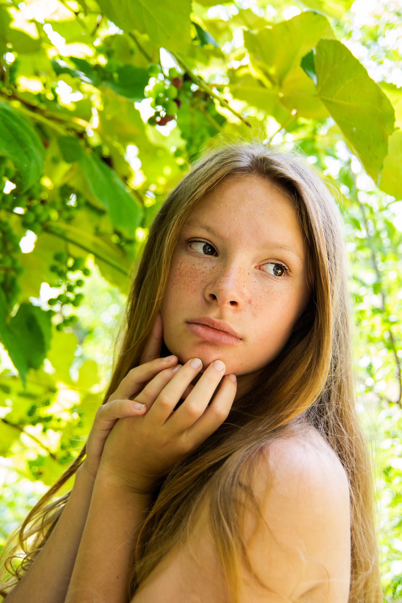 Portret młodej kobiety z piegami, zamyślona poza na tle zielonych liści i winogron w naturalnym świetle, delikatne dłonie przy twarzy, spokojny wyraz.