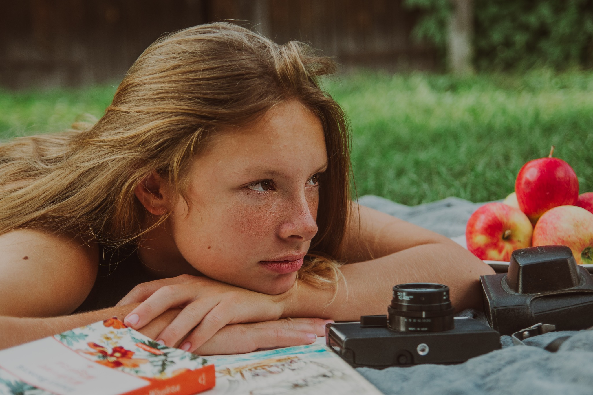 Młoda kobieta z piegami leży na kocu w plenerze, zamyślona, obok leżą jabłka, książka z motywem kwiatowym i stary aparat fotograficzny.