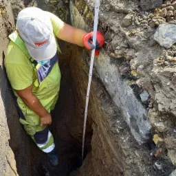 Pracownik w kombinezonie mierzy głębokość wykopu za pomocą składanej miarki, widoczne warstwy ziemi z kamieniami.