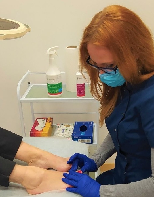 Kobieta w niebieskich rękawiczkach i masce higienicznej wykonuje zabieg na stopach klientki. Na stoliku obok widoczne środki dezynfekcyjne i chusteczki higieniczne.