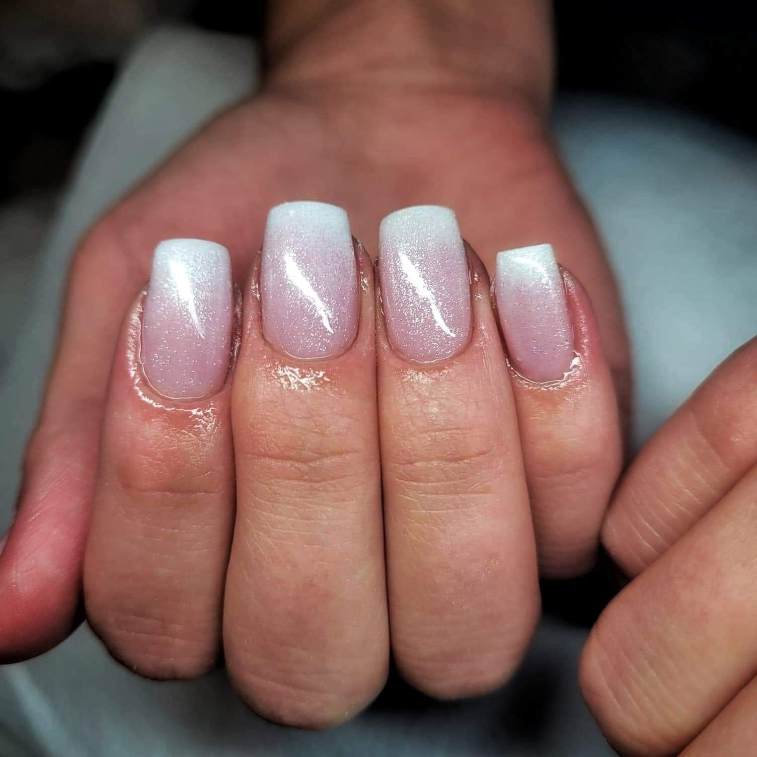 Dłoń z kwadratowymi paznokciami z efektem ombre od różowego do białego, wykończone błyszczącym lakierem.