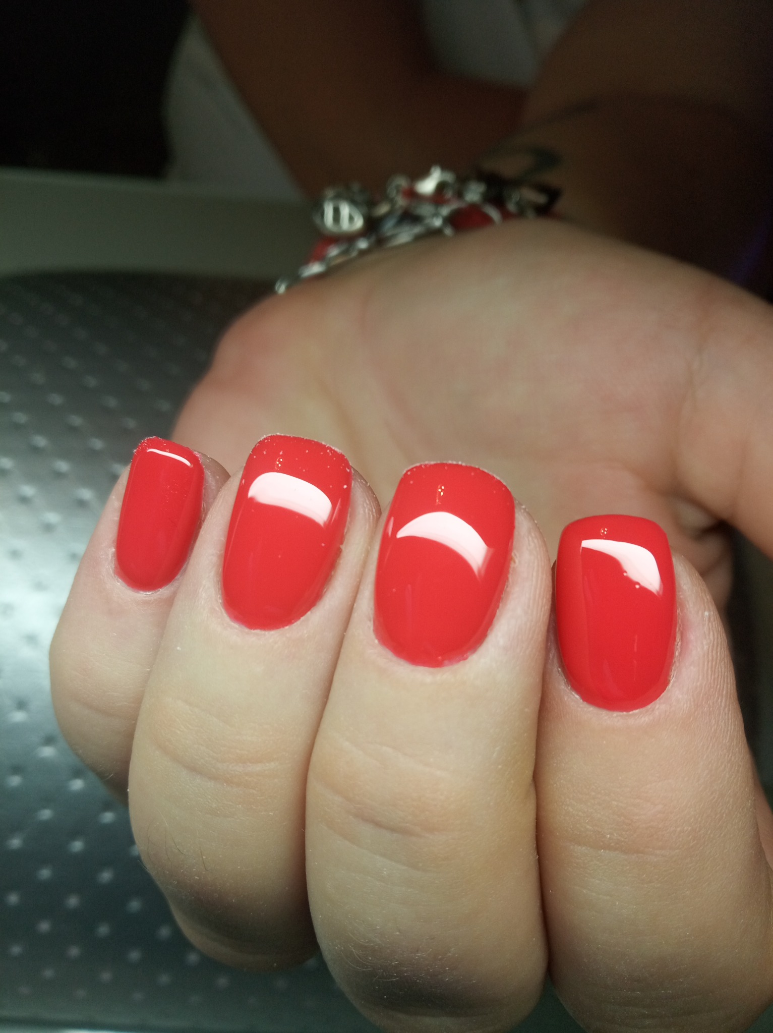 Dłoń z pomalowanymi na jaskrawy czerwony kolor paznokciami o kwadratowym kształcie, widoczny srebrny charms na nadgarstku