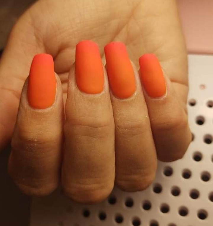 Dłoń z pomarańczowymi, matowymi paznokciami o prostokątnym kształcie, utwardzonymi pod lampą UV.