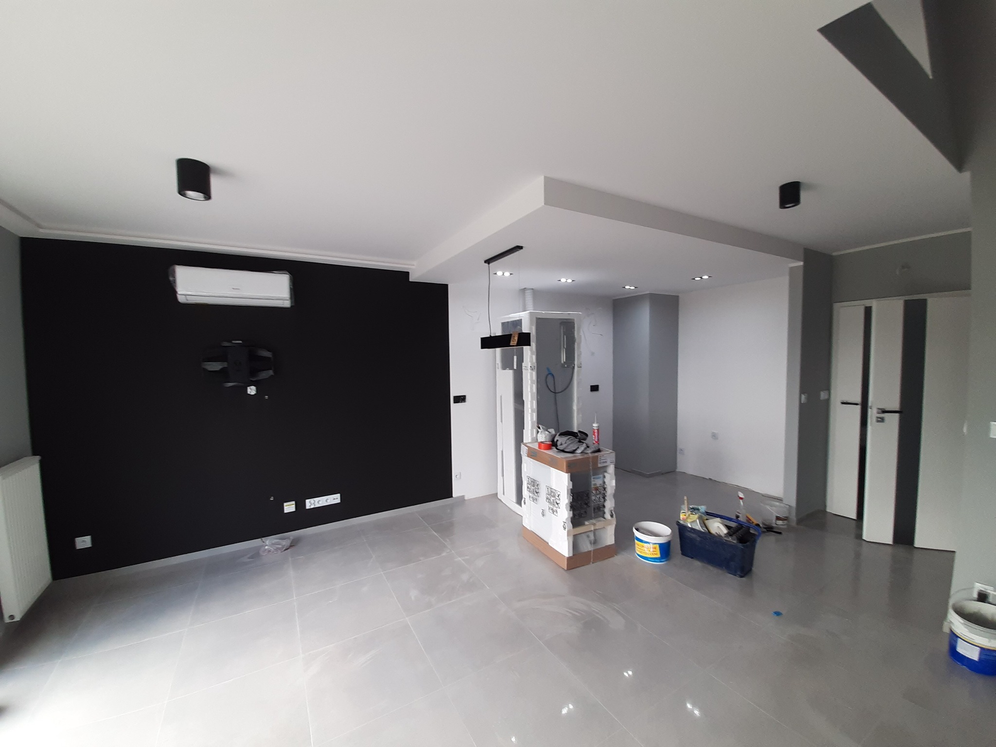 Wnętrze po częściowym remoncie: czarna ściana z klimatyzacją, biały sufit z punktowym oświetleniem, szare płytki podłogowe, narzędzia malarskie i wiadro z farbą w pomieszczeniu.