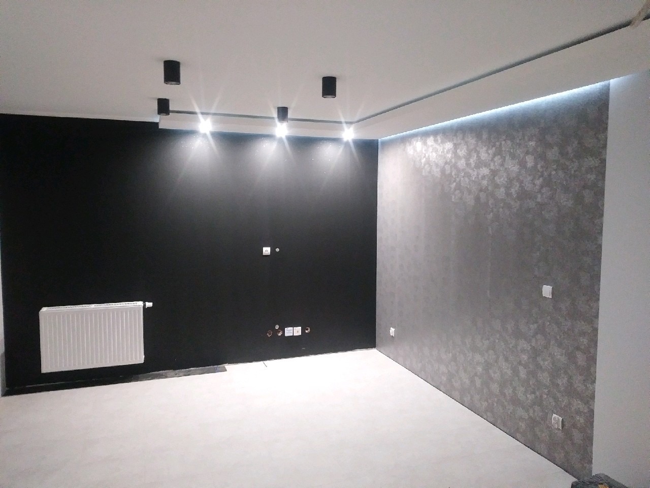 Wykończone wnętrze: czarna ściana z grzejnikiem, ściana z dekoracyjną tapetą w odcieniach szarości z podświetleniem LED przy suficie, biała podłoga.