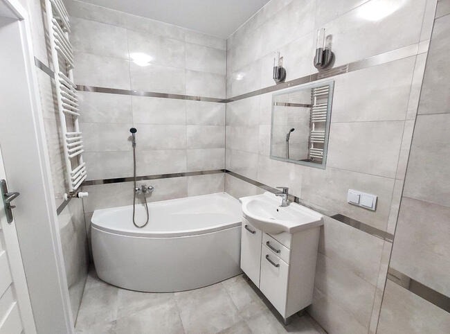 Wykończona łazienka w jasnych odcieniach szarości z narożną wanną, umywalką z szafką i grzejnikiem drabinkowym; dekoracyjne metalowe listwy akcentują płytki.