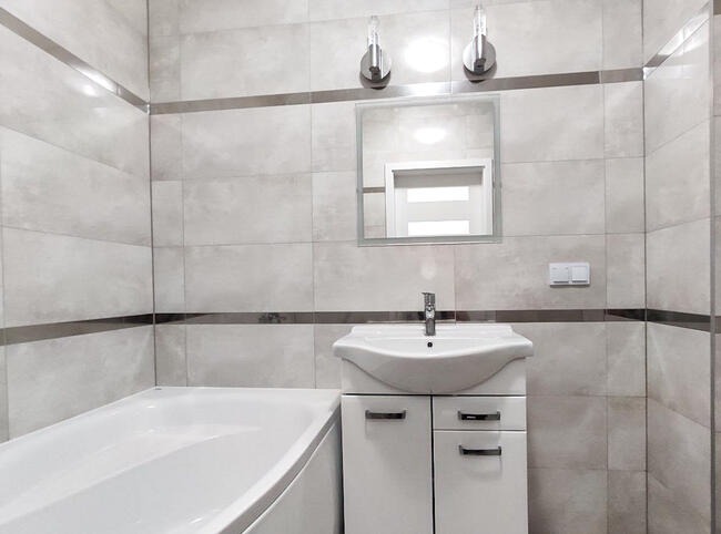 Jasna łazienka z wanną, umywalką z szafką i lustrem, wykończona płytkami w odcieniach szarości z dekoracyjnymi, poziomymi paskami.