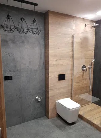 Nowoczesna łazienka z szarą i drewnopodobną okładziną, wisząca toaleta i kabina prysznicowa ze szklaną ścianką. Nad toaletą widoczny czarny przycisk spłuczki, a nad umywalką industrialna lampa...