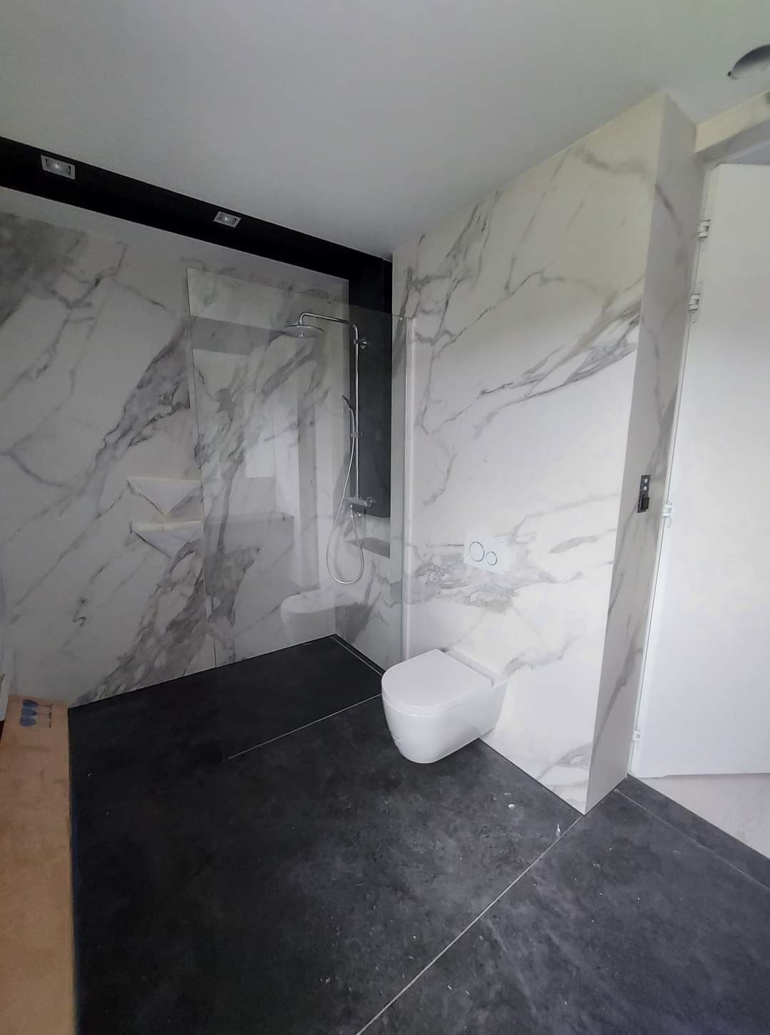 Nowoczesna łazienka z marmurowymi płytami na ścianach, kabiną prysznicową typu walk-in i podwieszaną toaletą, wykończona czarną podłogą.