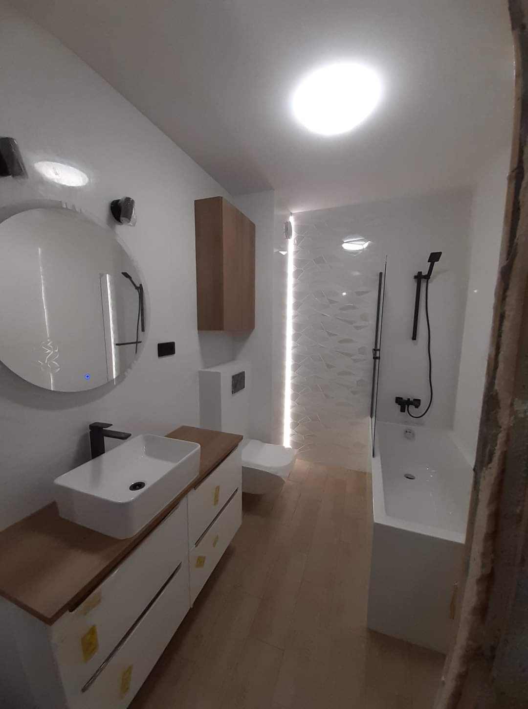 Nowoczesna łazienka z podświetlaną ścianą z dekoracyjną białą glazurą, okrągłym lustrem z podświetleniem LED, białą szafką pod umywalką z drewnianym blatem i czarną armaturą.