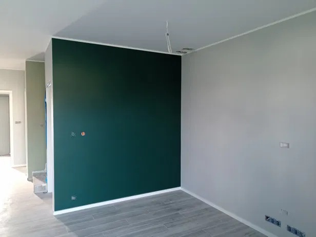 Pomieszczenie po malowaniu: ściana w kolorze butelkowej zieleni kontrastuje z szarą ścianą, widoczne gniazdka elektryczne i drewnopodobne panele na podłodze.