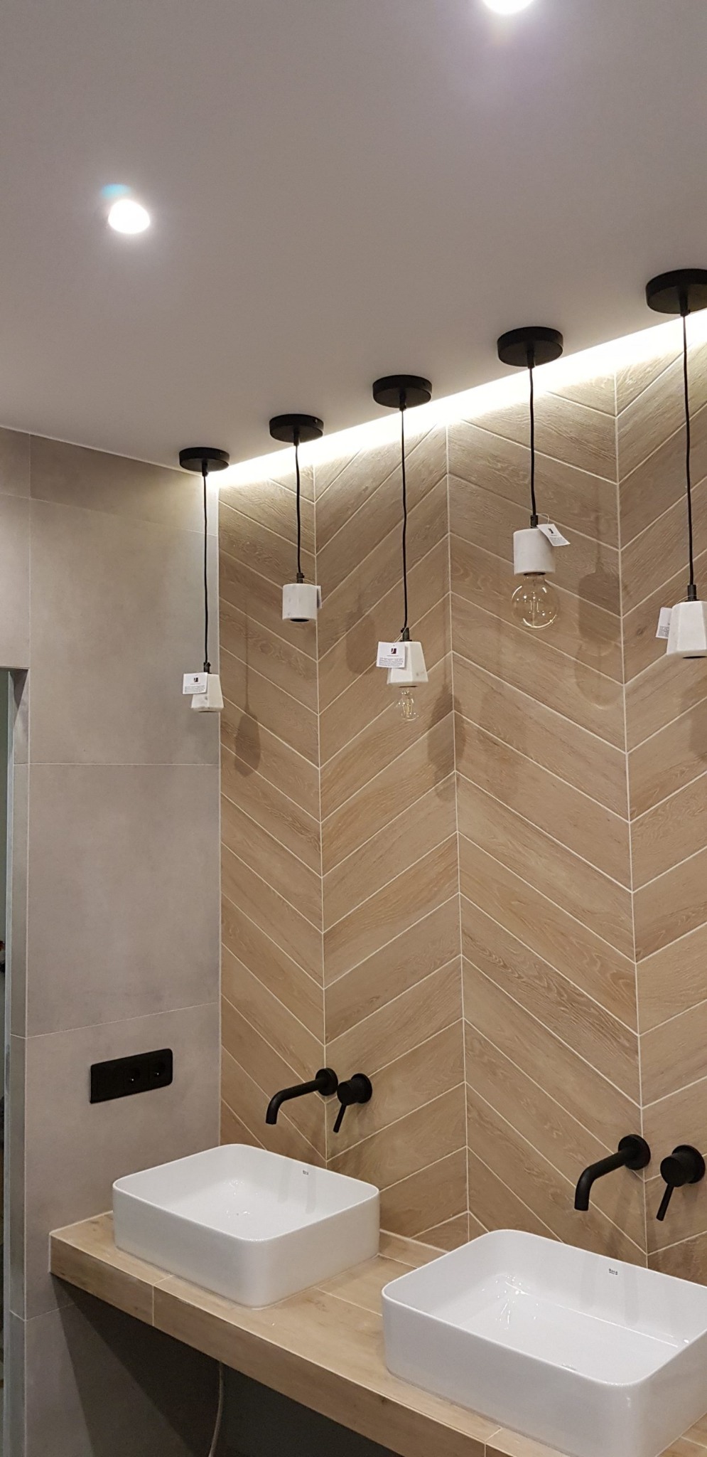 Nowoczesna łazienka z dwoma białymi umywalkami nablatowymi, czarnymi bateriami i dekoracyjną ścianą z płytek ułożonych w jodełkę; podwieszane lampy z marmurowymi elementami i żarówkami Edisona.