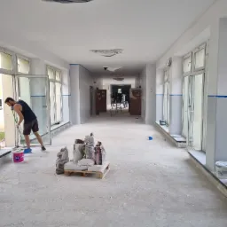 Korytarz w trakcie malowania: pracownik z wałkiem przy oknie, zabezpieczone lampy na suficie, worki z zaprawą na palecie, przygotowane ściany z niebieską taśmą malarską.