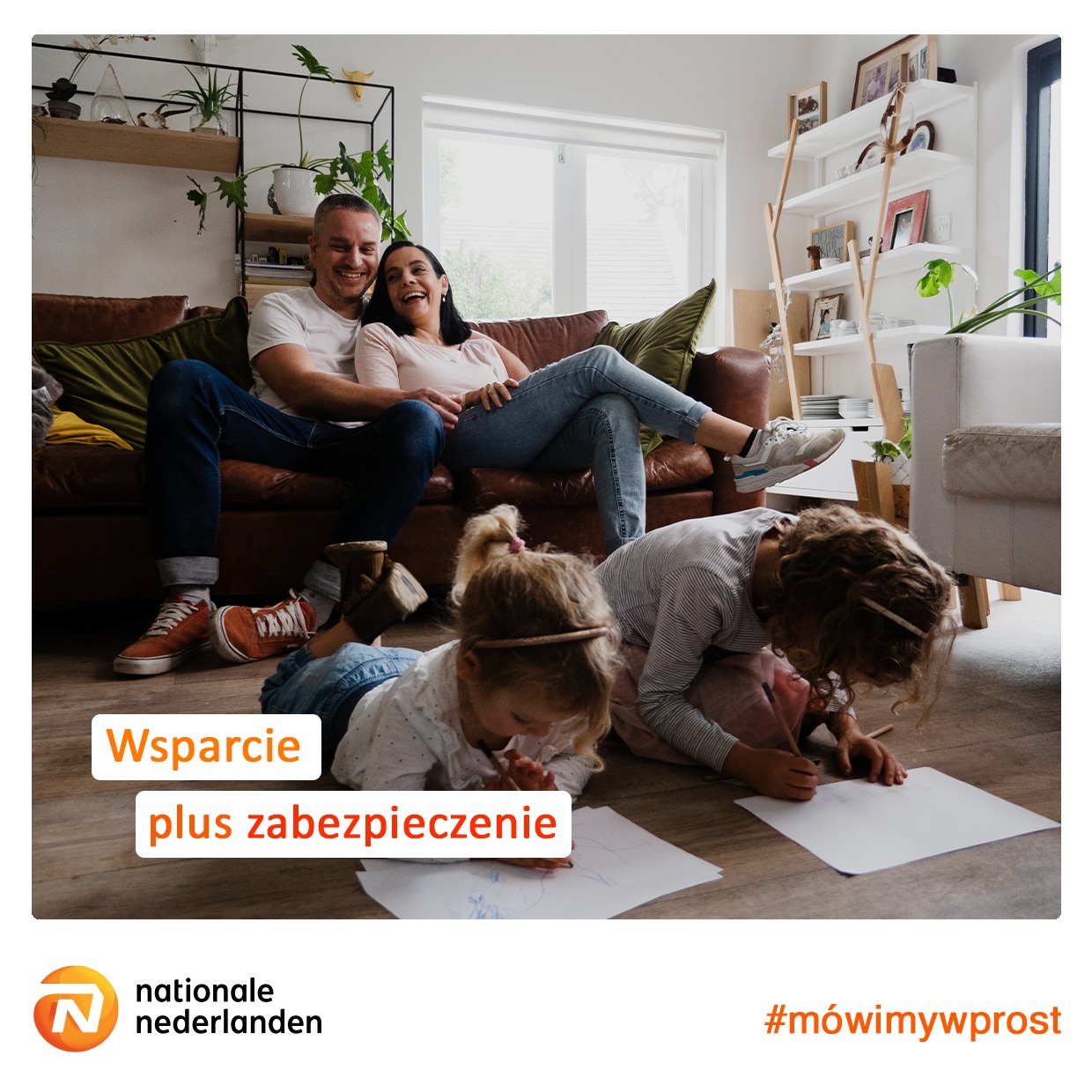 Uśmiechnięta rodzina: rodzice siedzą na skórzanej kanapie, a dwie córki leżą na podłodze rysując kredkami na papierze w jasnym, przestronnym salonie.