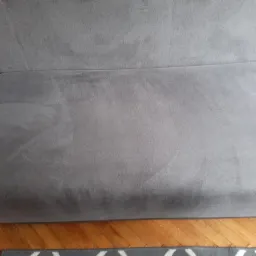 Szara kanapa z widocznymi śladami po czyszczeniu, częściowo widoczna podłoga drewniana i fragment dywanu w geometryczny wzór.