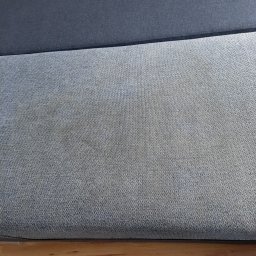 EMILIA MICHALCZYK - Fragment dwukolorowej tapicerki z widocznymi plamami na jaśniejszej części, leżącej na drewnianej podłodze.