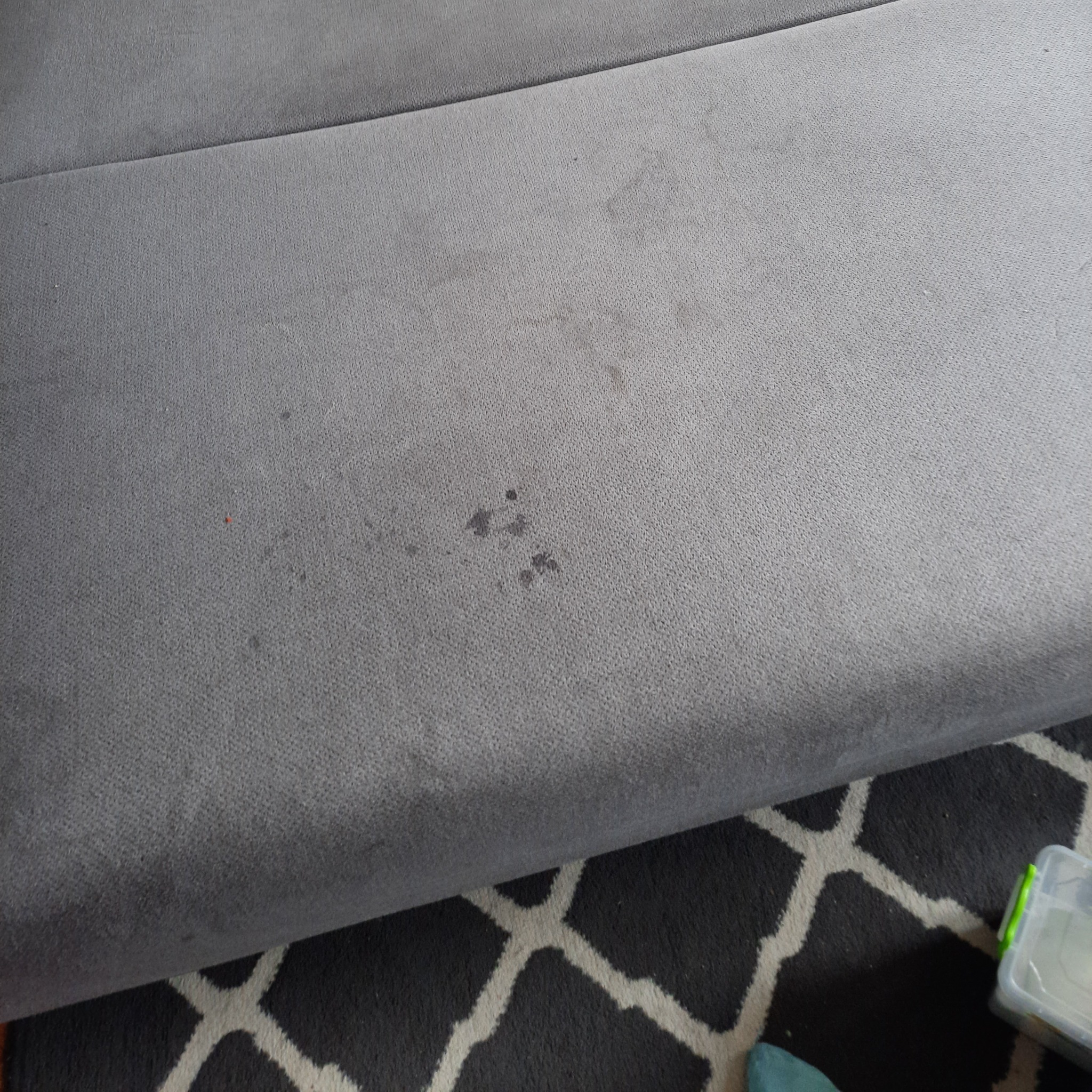 Fragment szarej, tapicerowanej kanapy z widocznymi plamami na siedzisku, leżącej na czarno-białym dywanie w geometryczny wzór.