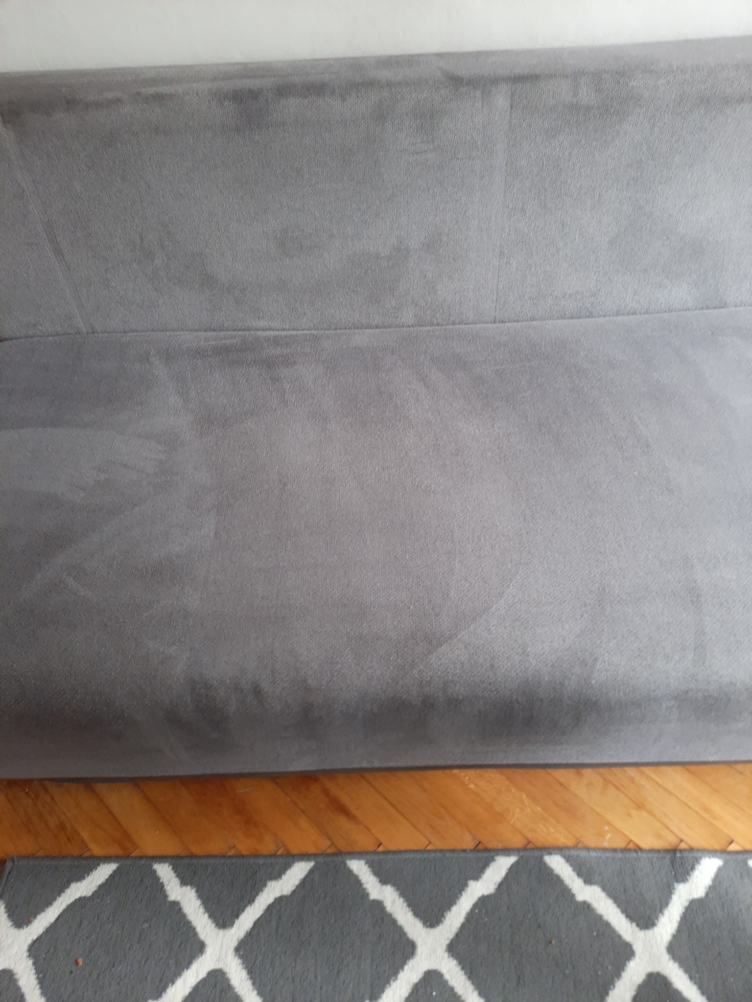 Szara kanapa z widocznymi śladami po czyszczeniu, częściowo widoczna podłoga drewniana i fragment dywanu w geometryczny wzór.