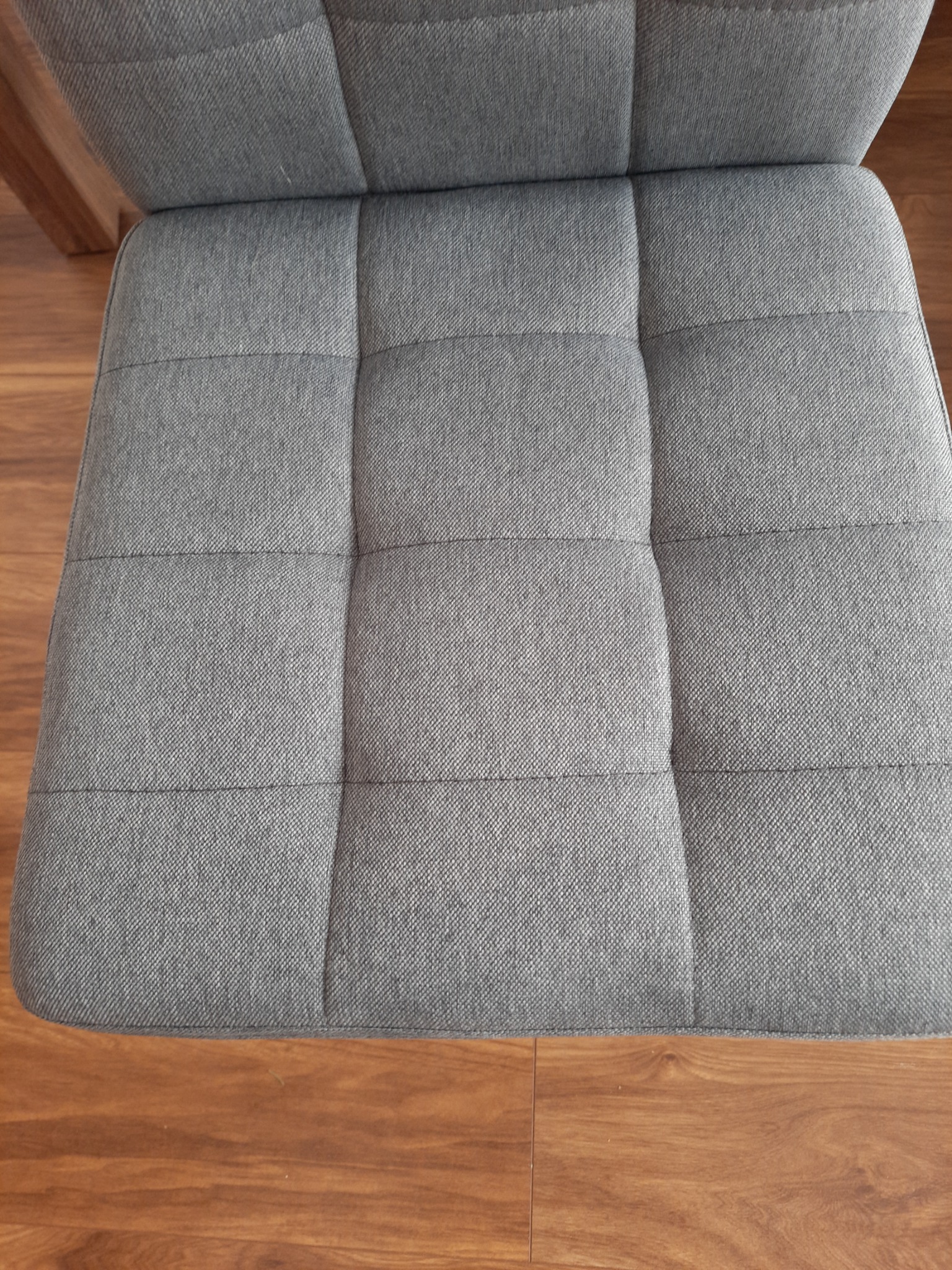 Fragment szarego, tapicerowanego siedziska krzesła lub fotela, widok z góry, na tle drewnianej podłogi.