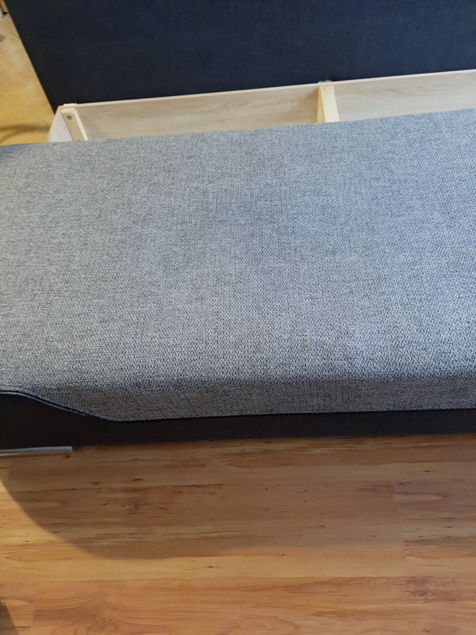 Szary tapicerowany element sofy z widocznym drewnianym stelażem i fragmentem podłogi z paneli