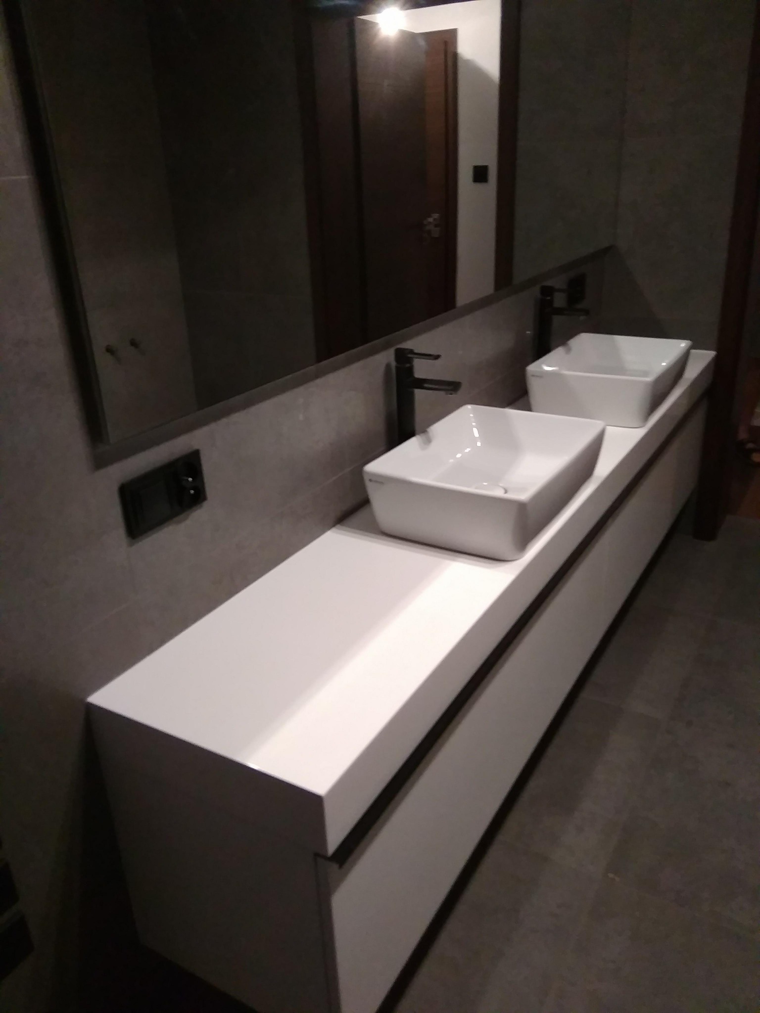 Nowoczesna łazienka z dwiema białymi umywalkami nablatowymi o kwadratowym kształcie, zamontowanymi na długim, białym blacie z szafką, czarne baterie, szara ściana wykończona płytkami, widoczne...